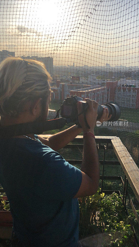 一个拿着数码单反相机的男人，自由摄影，以公寓/公寓大楼为背景，站在公寓阳台上用变焦镜头拍照，一个有着漂白金发的男人，看着相机屏幕的取景器。